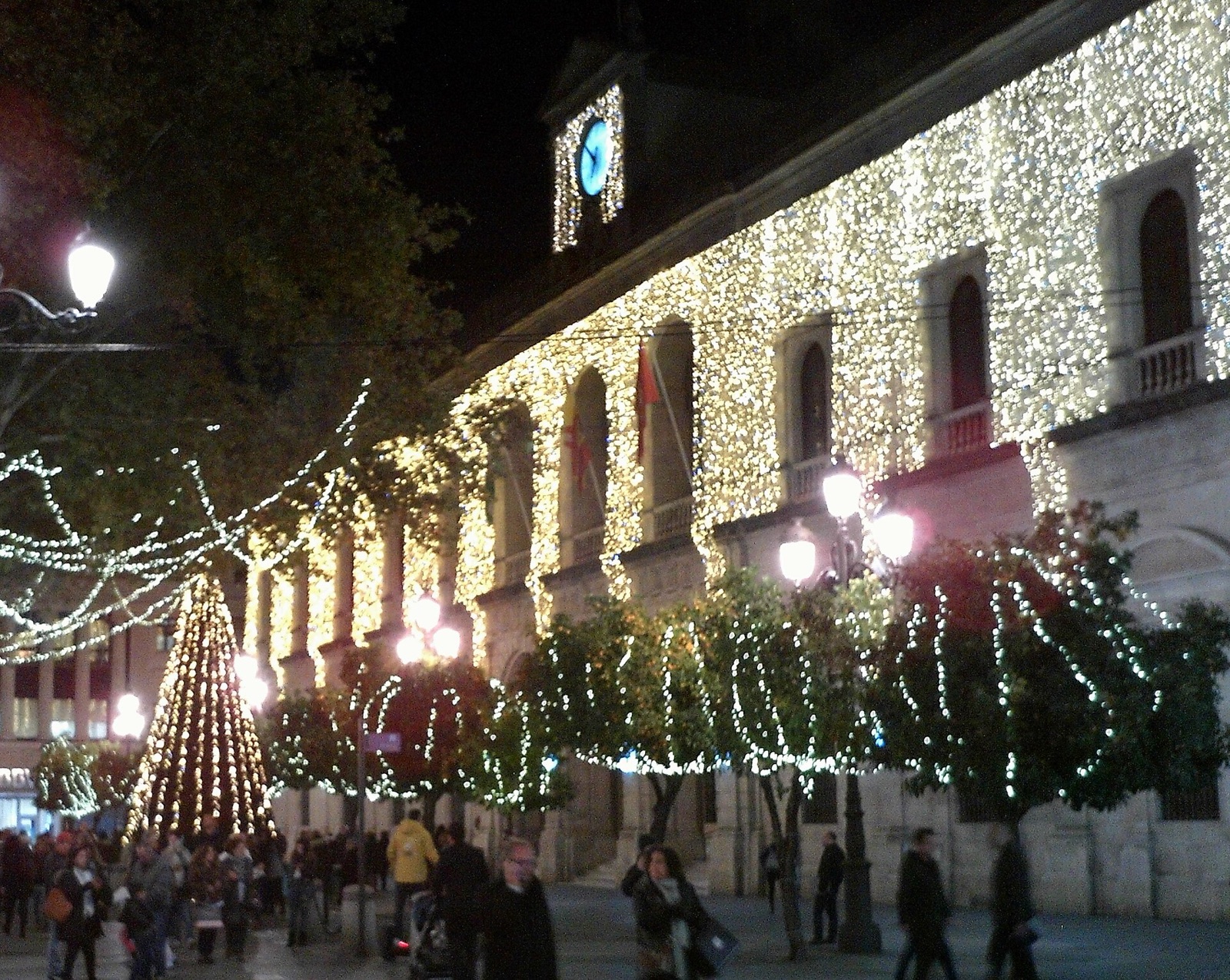 Fachada del Ayuntamiento de Sevilla cubierta de luces de Navidad, con un árbol luminoso y paseantes