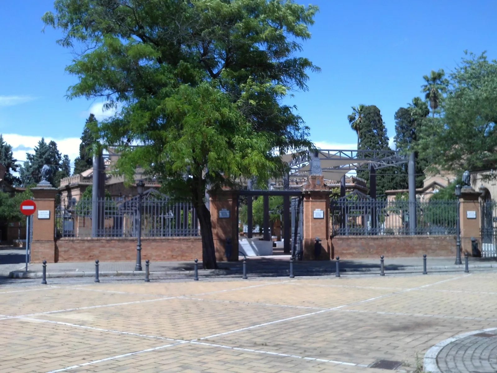 Entrada principal del cementerio de San Fernando de Sevilla, con verja de hierro entre pilares de ladrillo y un gran árbol delante, bajo un cielo azul