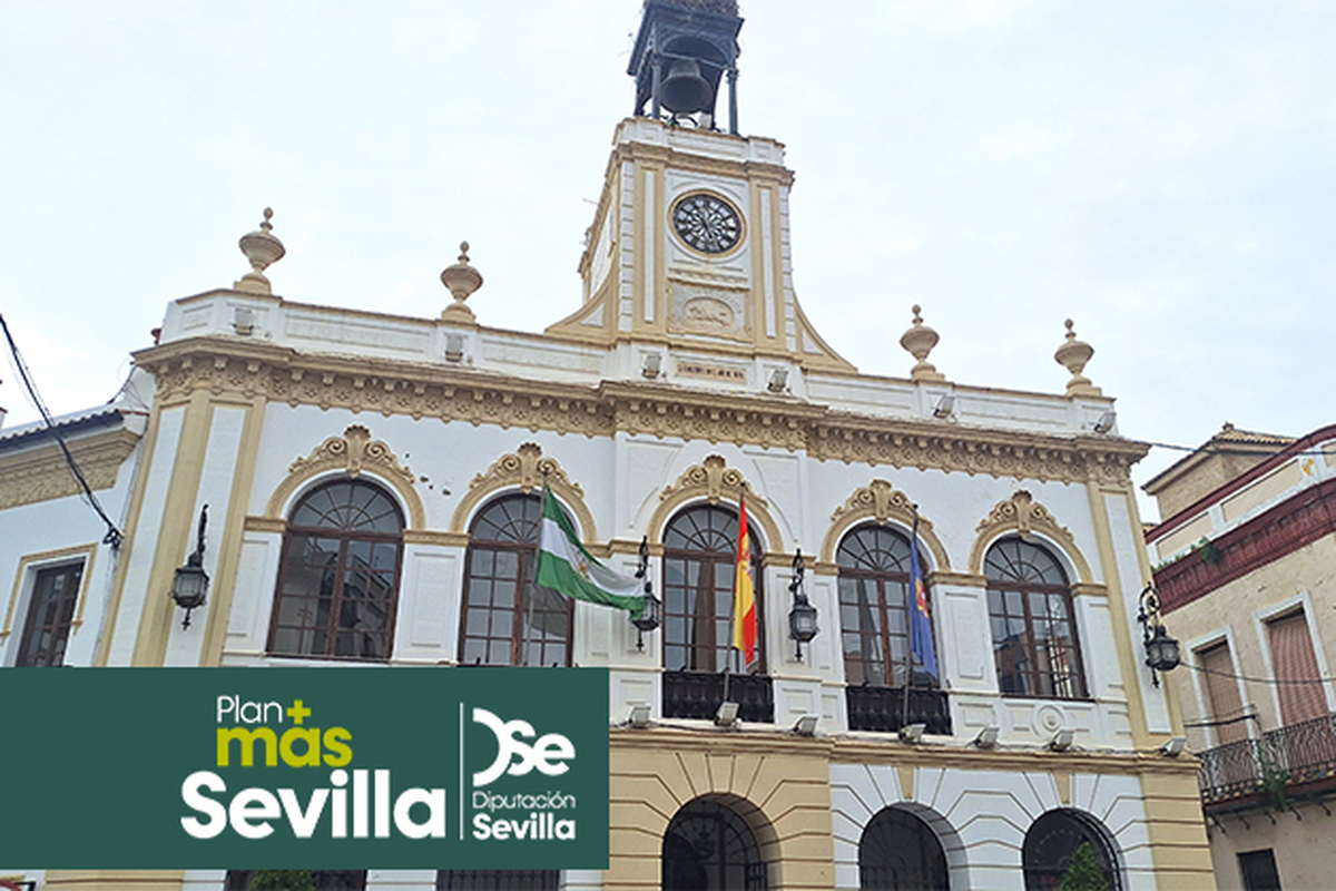 Edificio municipal blanco y amarillo con torre y reloj central, arcos y balcones con banderas, y un rótulo verde de Sevilla abajo.