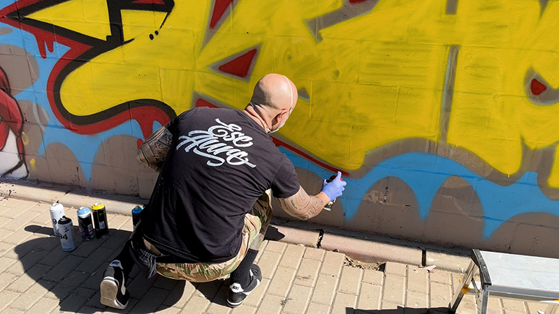 Un hombre rapado con camiseta negra pinta un grafiti amarillo y azul en una pared, arrodillado junto a botes de spray.