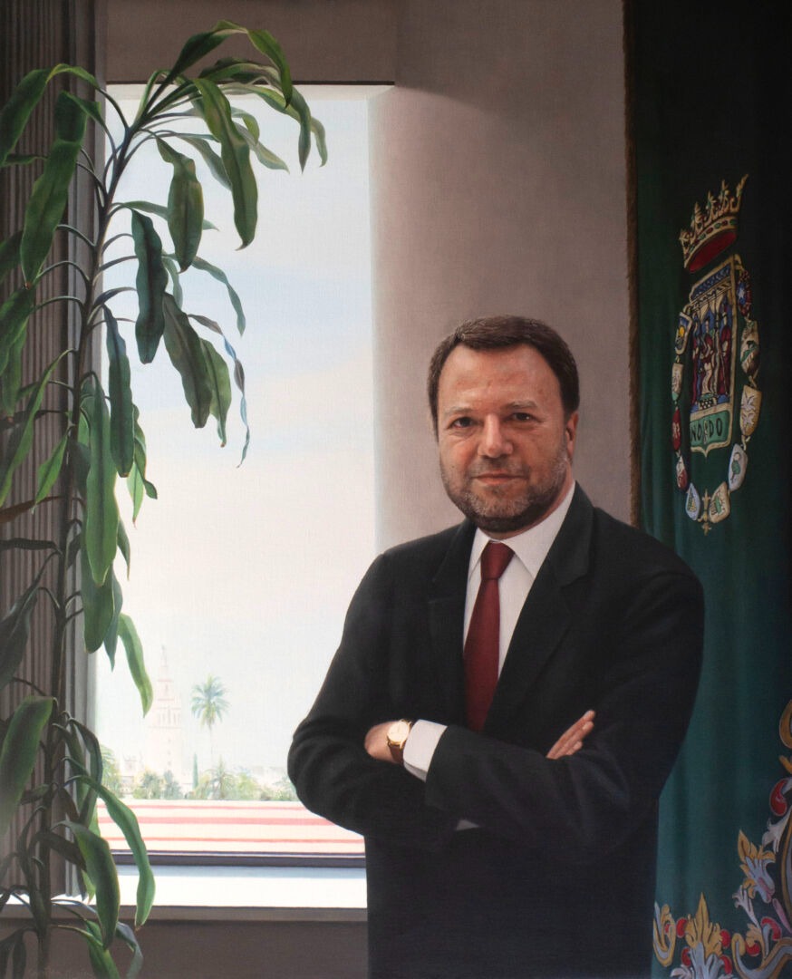 Hombre trajeado con corbata roja y brazos cruzados, junto a una ventana con una planta y una bandera verde con escudo.