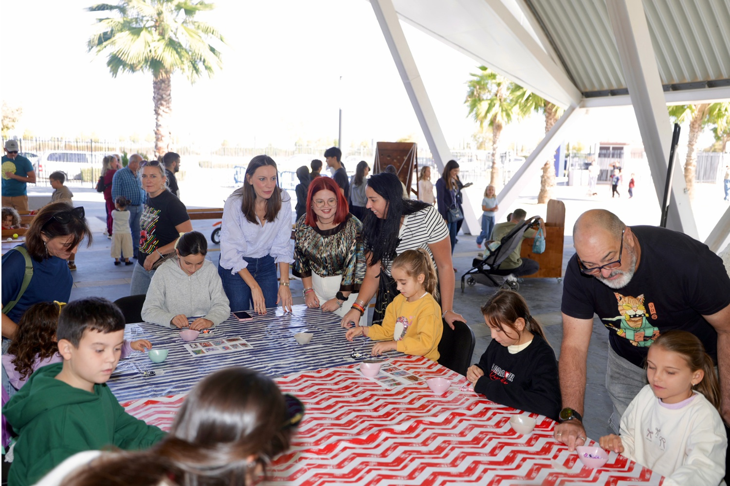 Niños y adultos alrededor de una mesa con mantel de rayas rojas y blancas, haciendo manualidades bajo una estructura con palmeras al fondo.