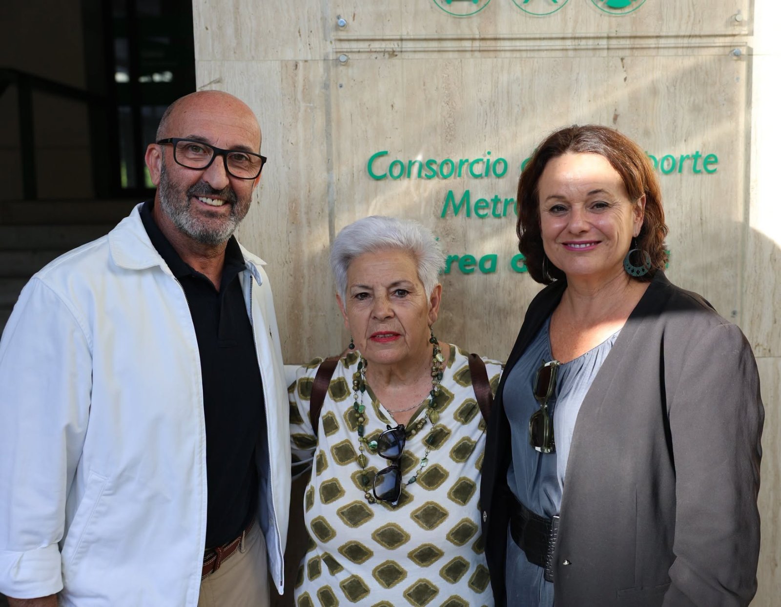 Tres personas adultas posan sonrientes ante un panel de piedra con el rótulo verde Consorcio de Transporte Metro.