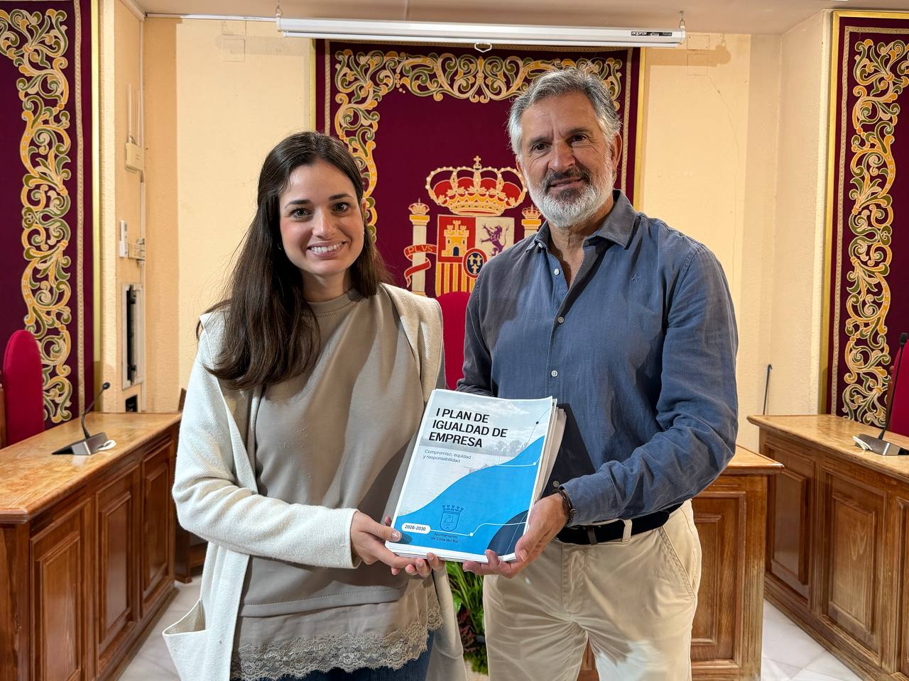 Una mujer y un hombre sostienen juntos un documento titulado I Plan de Igualdad de Empresa en una sala con escudo y cortinajes.