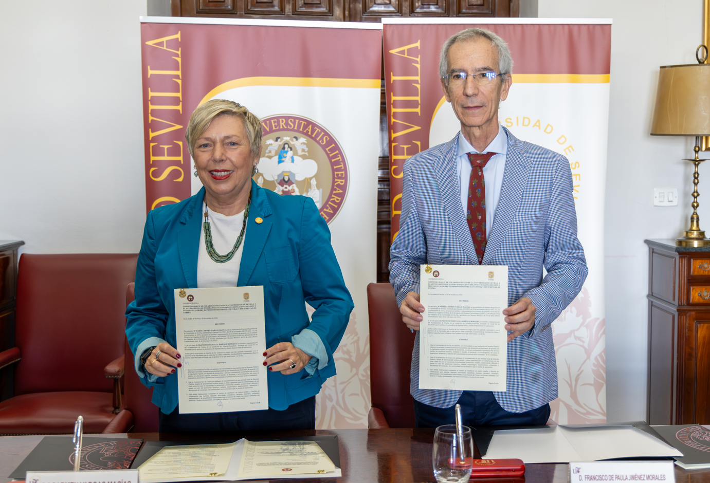 Una mujer con chaqueta azul y un hombre con americana de cuadros sostienen sendos documentos delante de dos carteles de la Universidad de Sevilla.