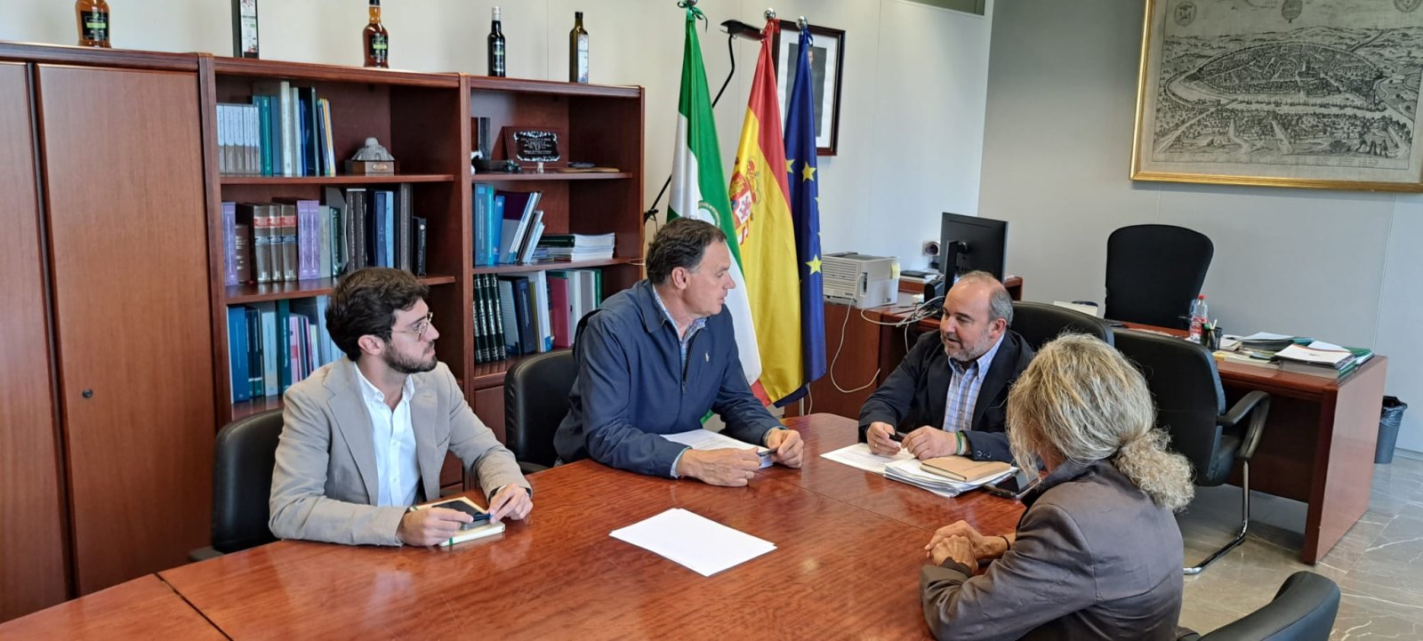 Cuatro personas conversando sentadas alrededor de una mesa de madera en un despacho con estanterías y banderas.
