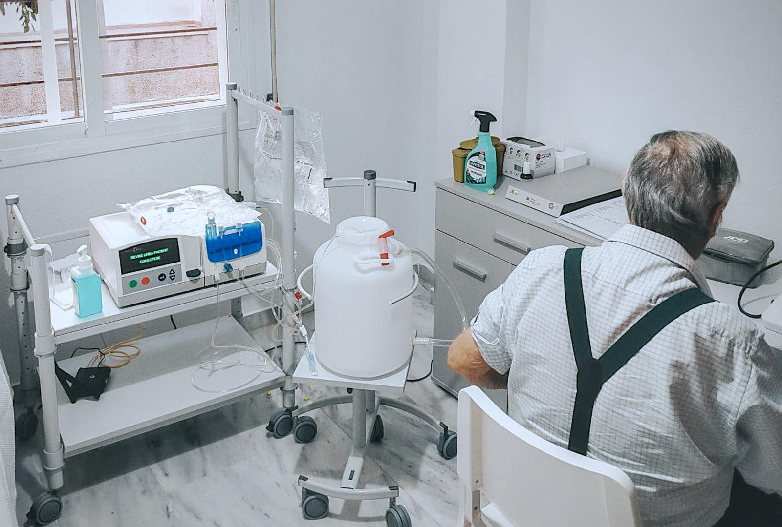 Un hombre mayor de espaldas, sentado junto a una máquina médica con bolsas y un bidón blanco en una habitación.