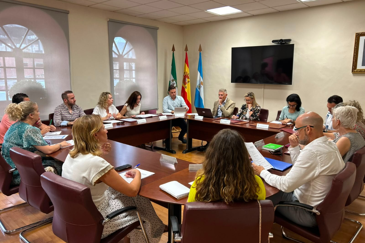 Una docena de personas sentadas alrededor de una mesa de reuniones ovalada, con portátiles y documentos, en una sala con banderas y una pantalla.