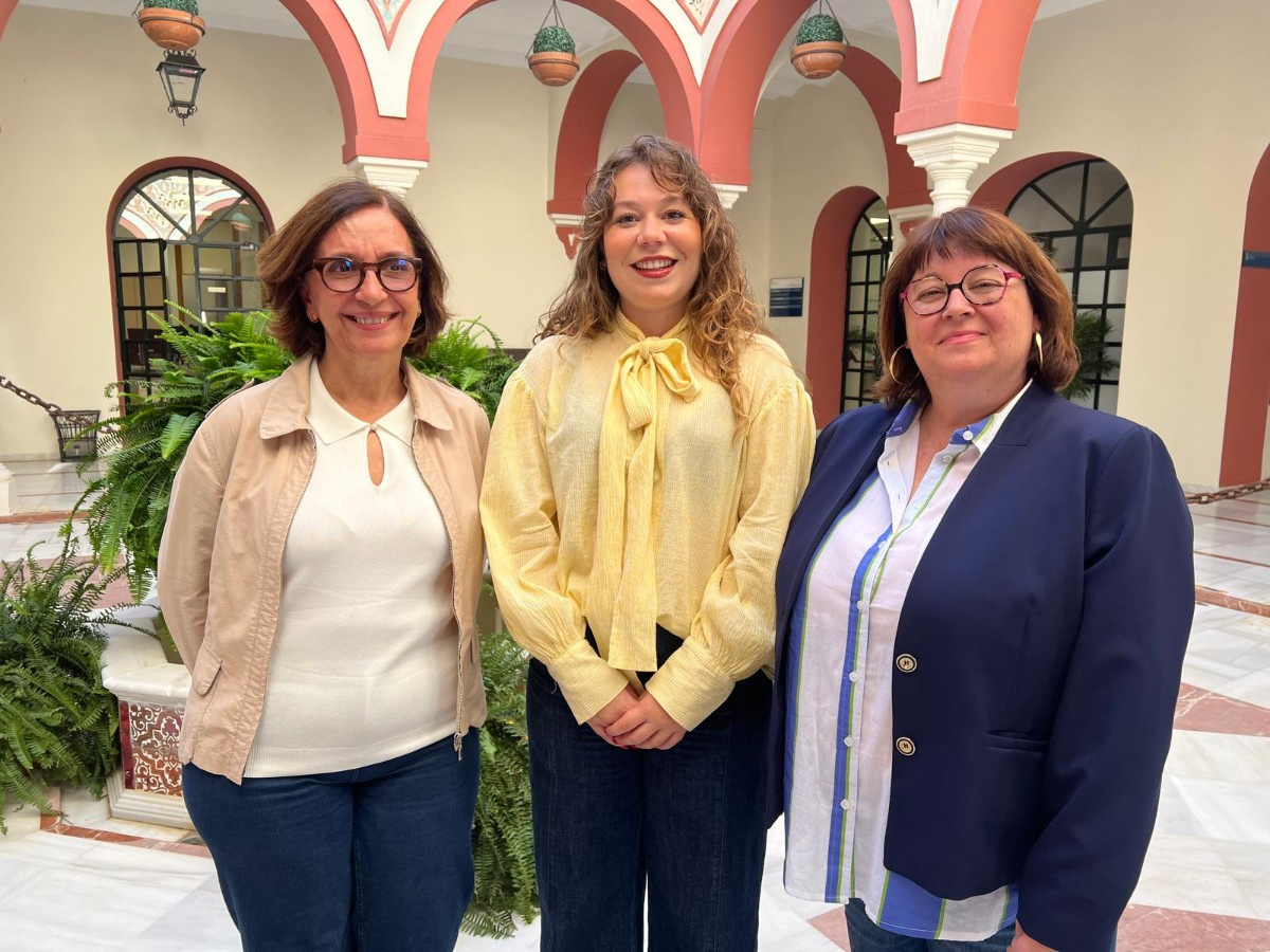 Tres mujeres de pie, sonriendo, en un patio con arcos rojiblancos y plantas.