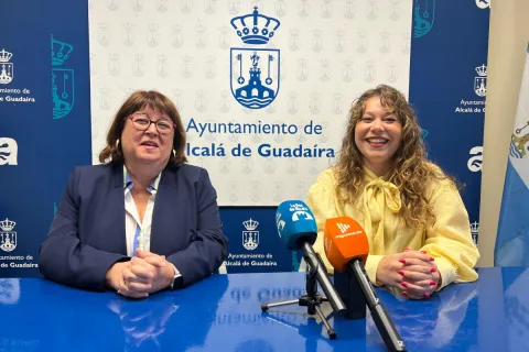 Dos mujeres sonríen sentadas ante una mesa azul con micrófonos, delante de un panel del Ayuntamiento de Alcalá de Guadaíra.