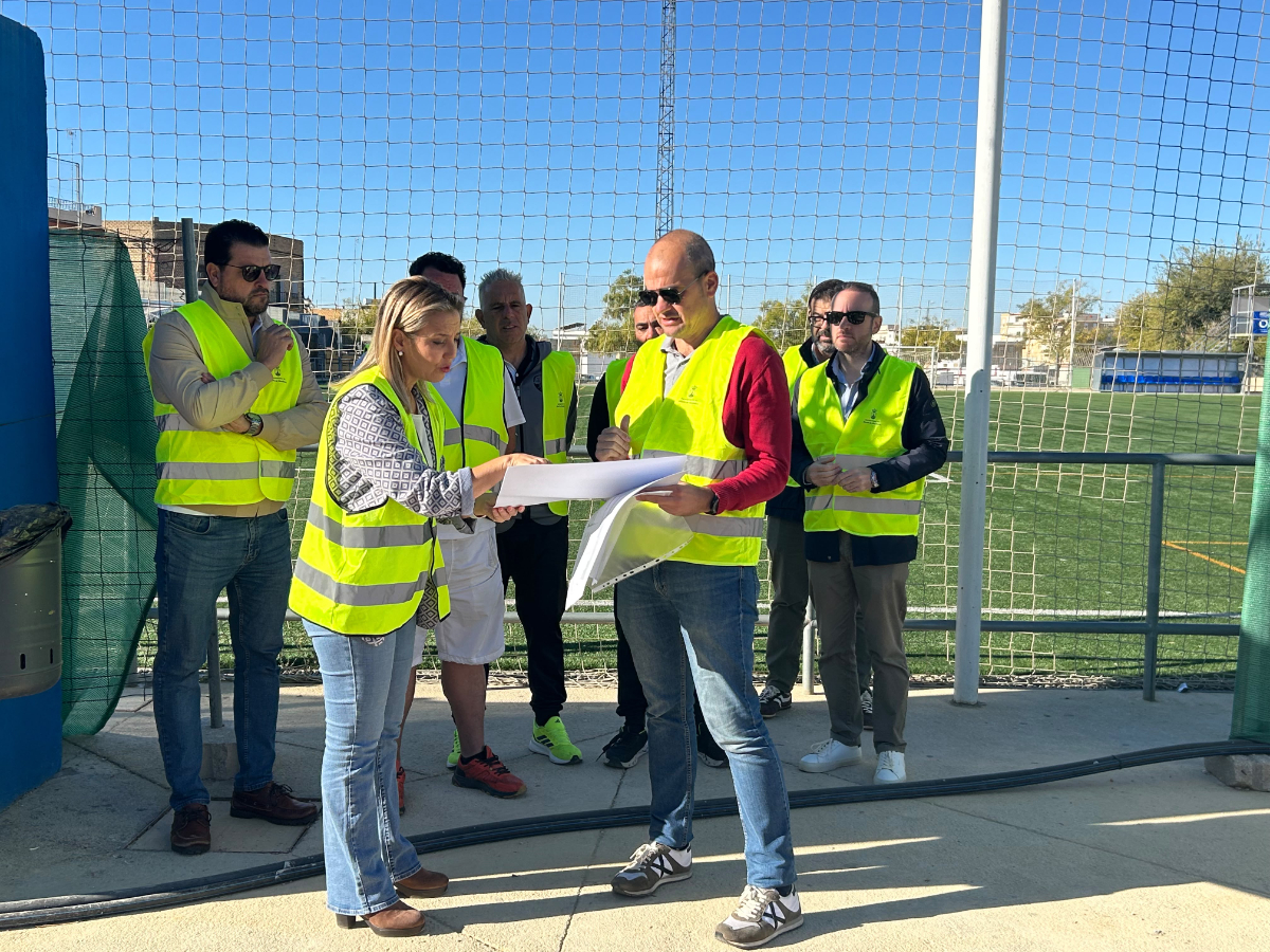 Un grupo de personas con chalecos reflectantes revisa unos planos junto a la valla de un campo de fútbol.