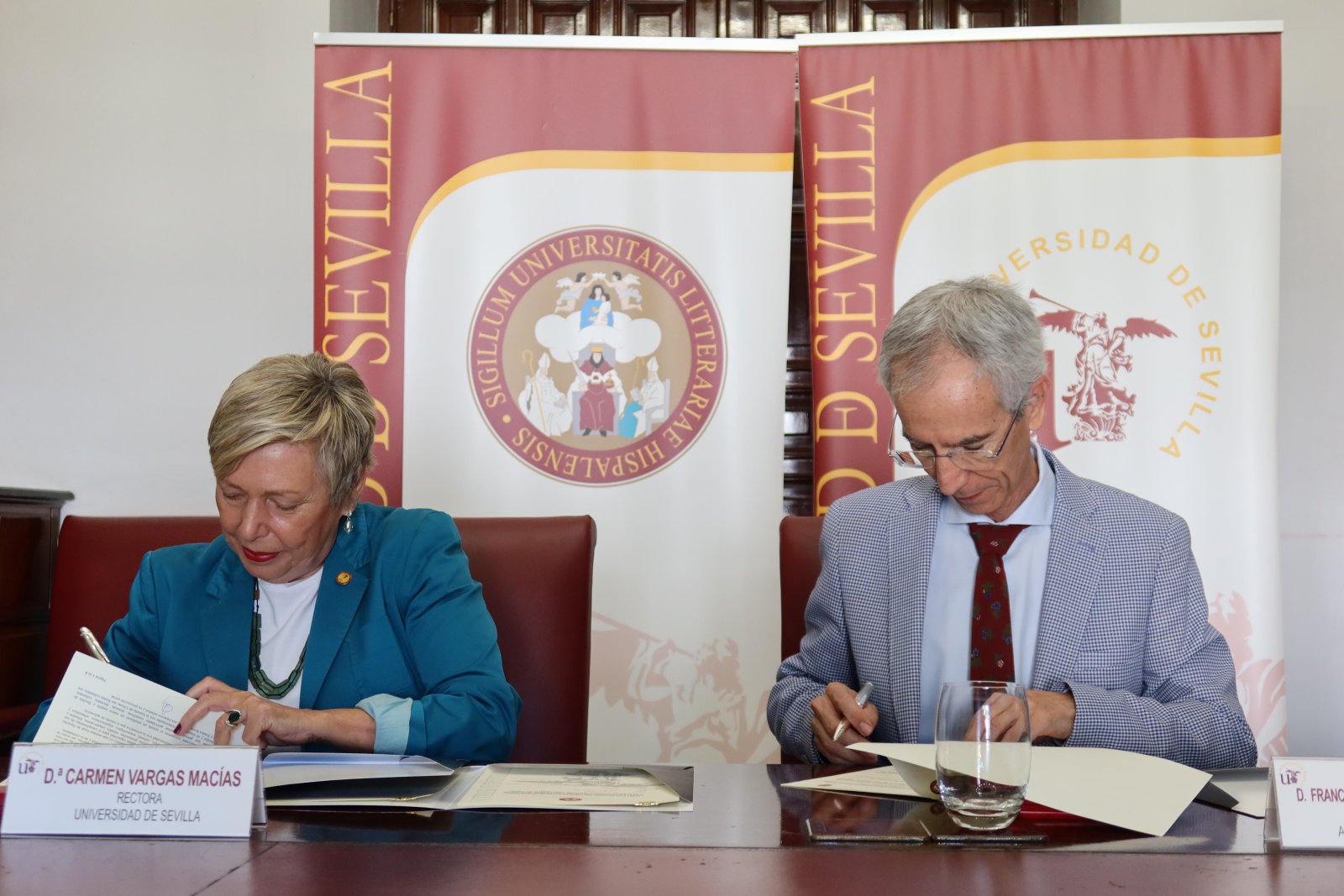 Una mujer y un hombre sentados ante una mesa firman documentos, con carteles de la Universidad de Sevilla al fondo.