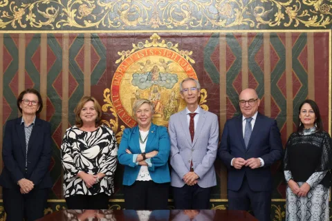 Seis personas trajeadas posan de pie ante un tapiz con un gran escudo bordado en dorado.