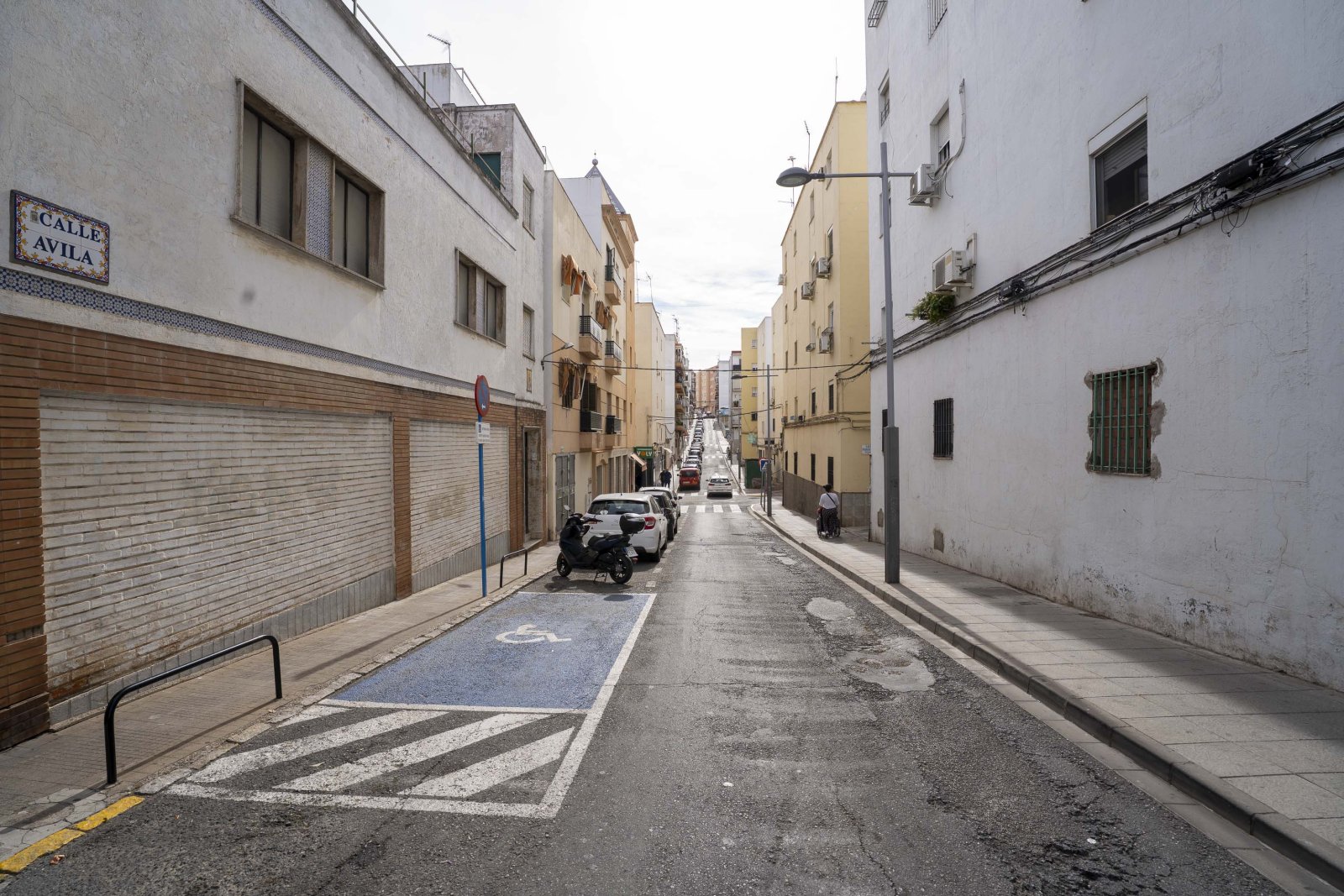 Calle estrecha de edificios blancos y amarillos con coches aparcados, una plaza azul de minusválidos y una placa que dice Calle Ávila.