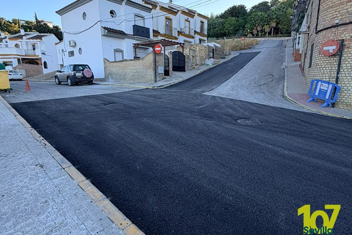 Calle recién asfaltada en cuesta con casas blancas a la izquierda, un todoterreno aparcado y una valla azul a la derecha.