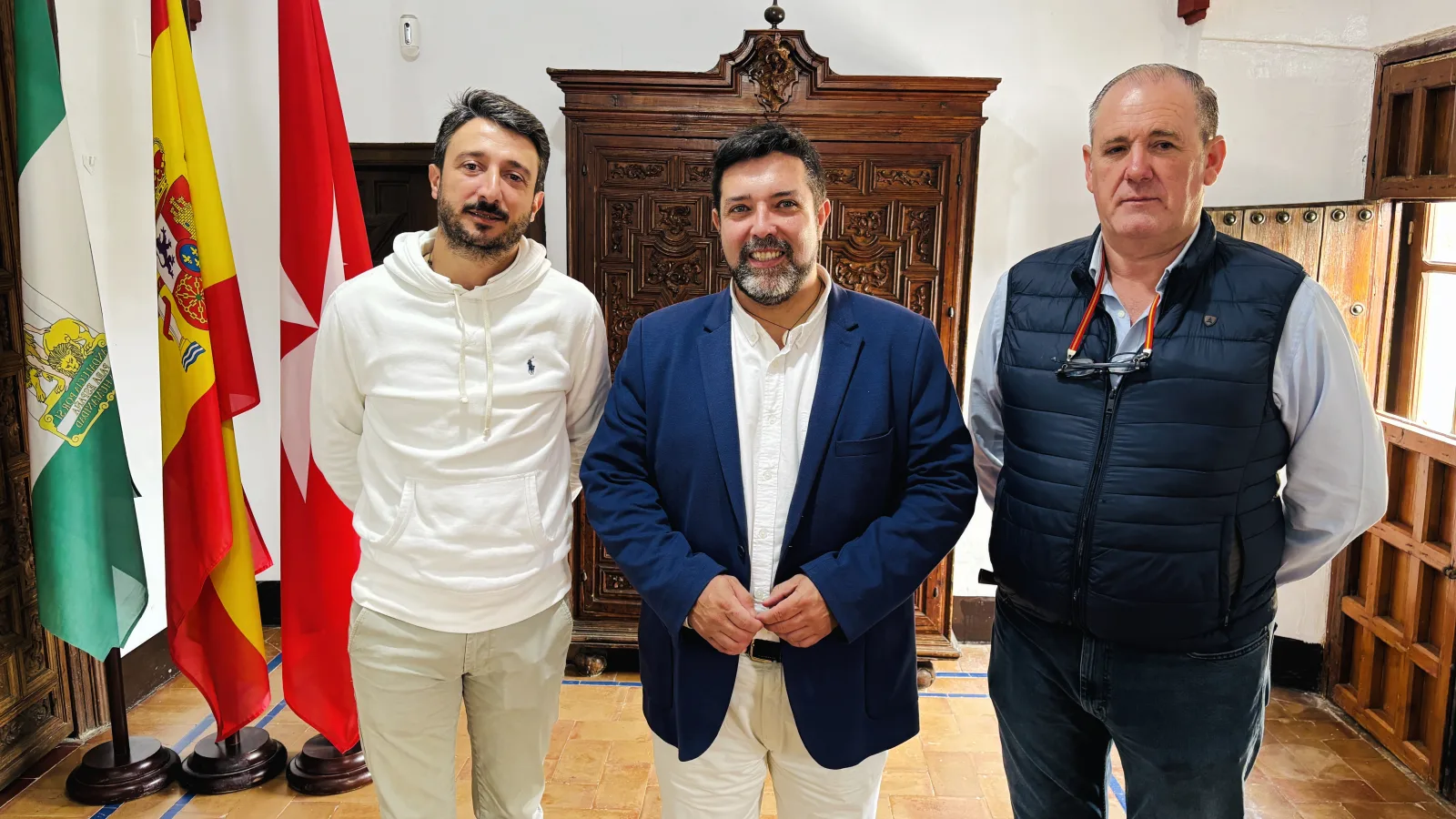 Tres hombres de pie posan en una sala con banderas de Andalucía y España y un armario de madera tallada al fondo.