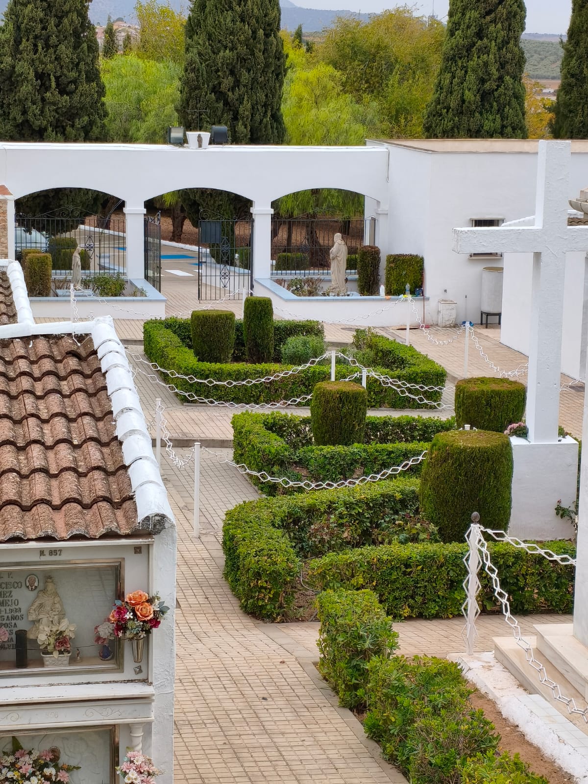 Nicho con flores y cruz blanca en un cementerio ajardinado con setos, arcos y cipreses al fondo.