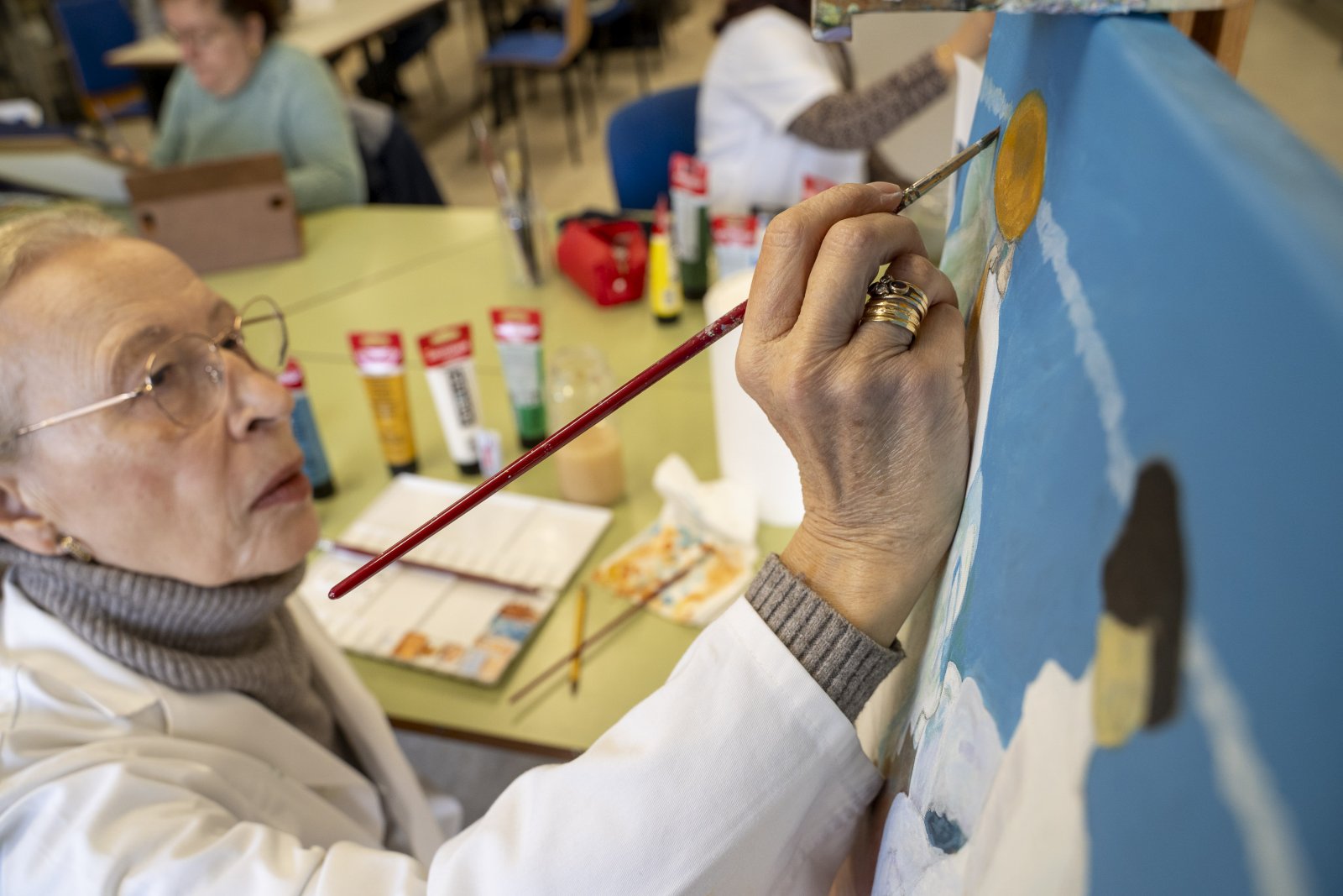 Una mujer mayor con gafas pinta un sol amarillo en un lienzo azul con un pincel, en una sala con más personas y tubos de pintura.