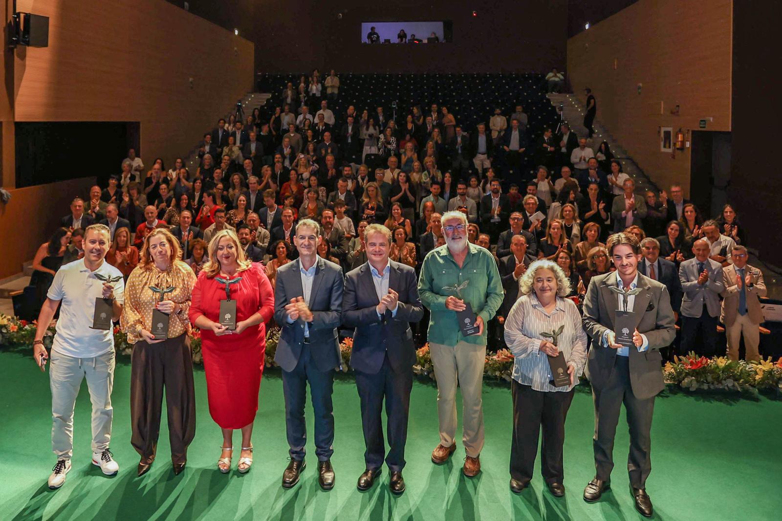 Ocho personas de pie sobre una alfombra verde sostienen trofeos ante un público sentado en un auditorio.