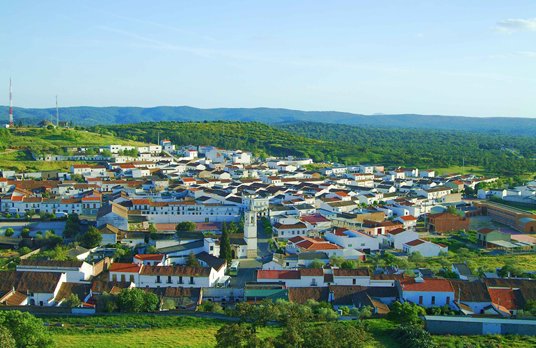 Vista panorámica de un pueblo de casas blancas con tejados rojizos, una torre de iglesia y colinas verdes al fondo.