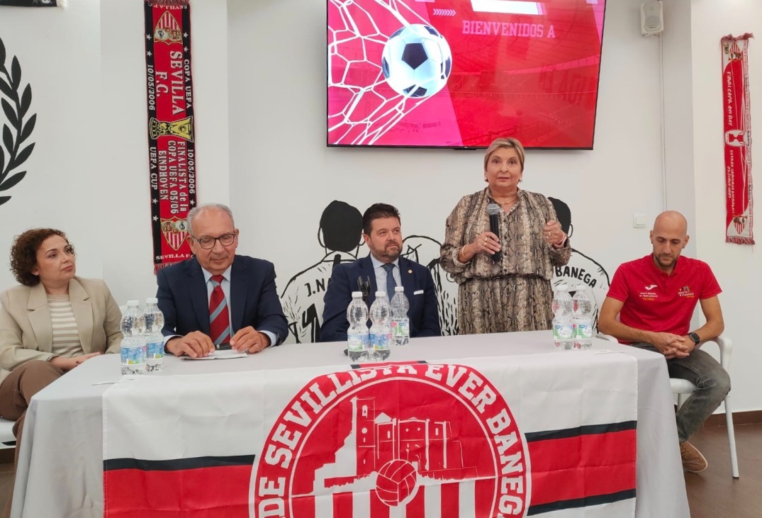 Cuatro personas sentadas tras una mesa con mantel del Sevilla FC y una mujer de pie hablando con un micrófono.