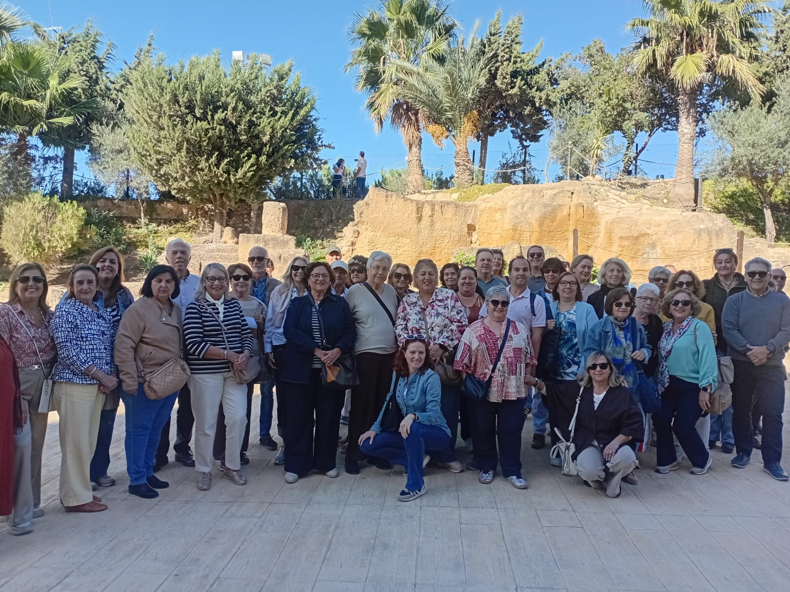 Un grupo numeroso de personas adultas posa de pie al aire libre ante un muro de piedra y palmeras.