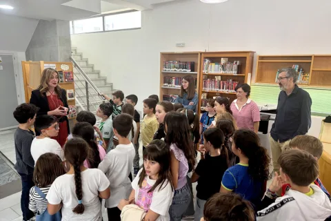 Una mujer adulta habla a un grupo de niños y niñas reunidos junto a estanterías con libros, en el interior de una biblioteca.