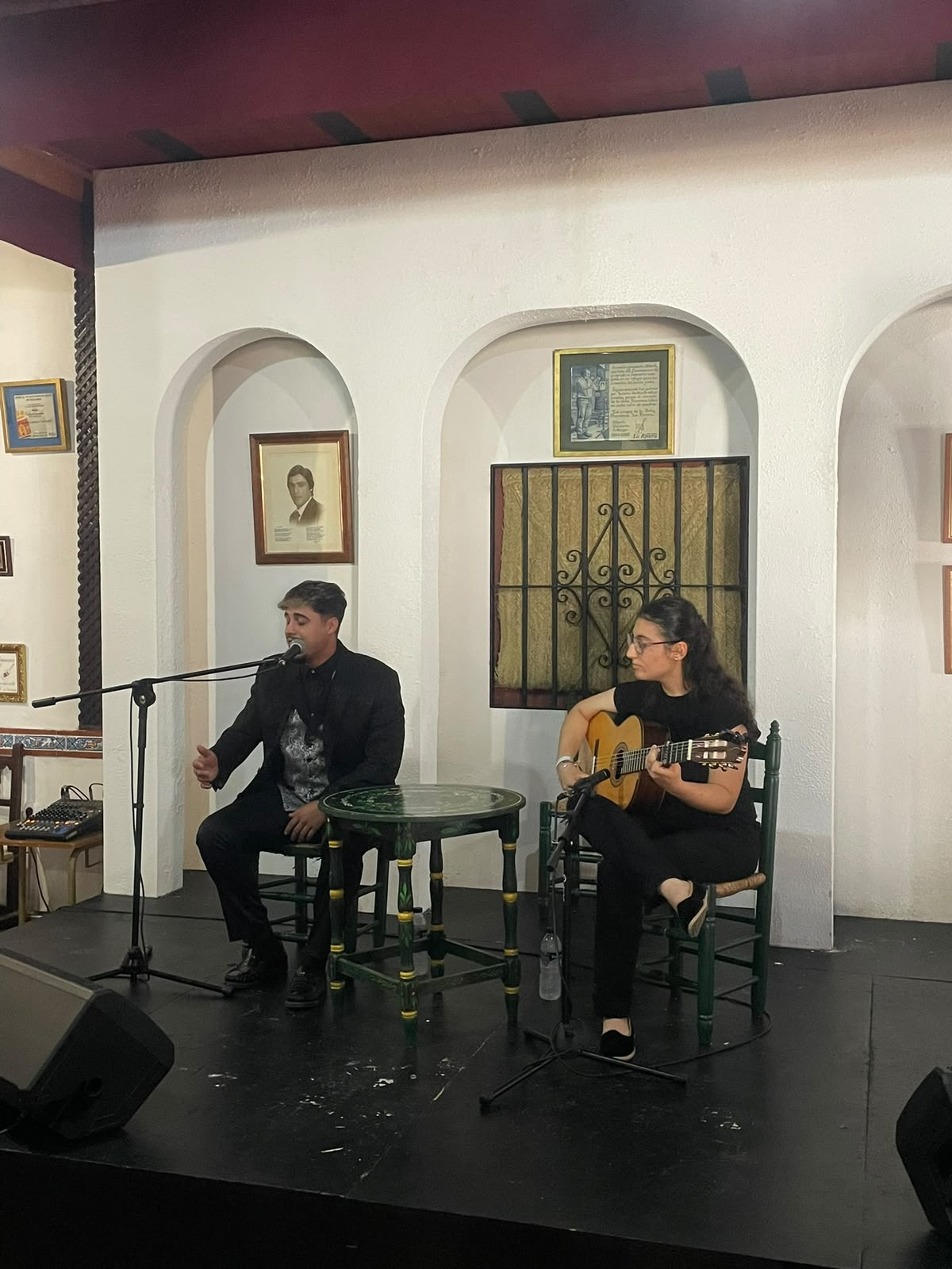 Un hombre canta ante un micrófono y una mujer toca la guitarra sentados en un escenario con arcos blancos y retratos enmarcados.