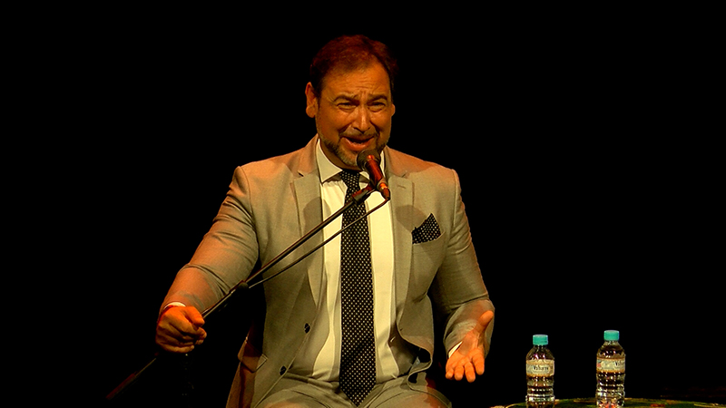 Hombre trajeado con corbata de lunares canta o habla ante un micrófono sobre un escenario oscuro, con dos botellas de agua delante.