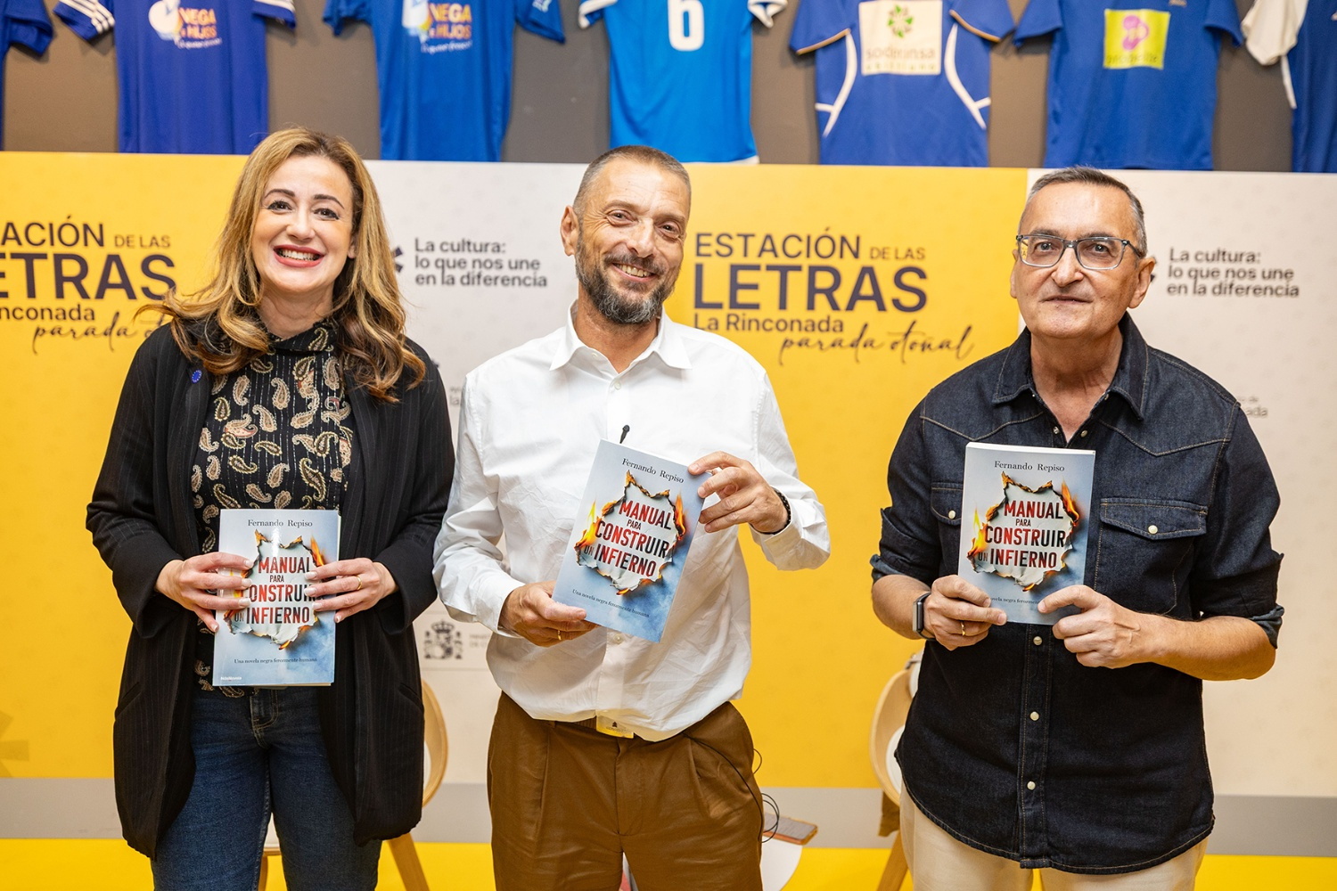Tres personas de pie sostienen el libro Manual para construir un infierno ante un panel amarillo con camisetas azules al fondo.