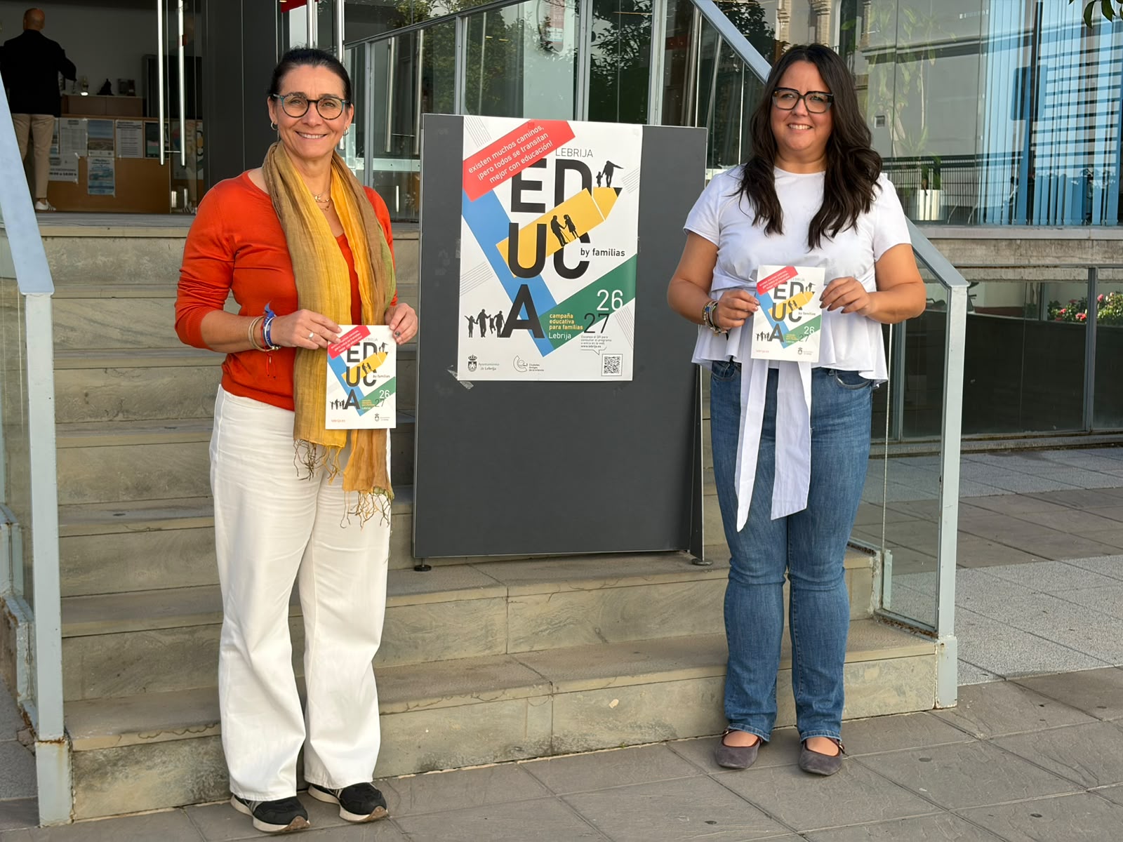 Dos mujeres de pie en unas escaleras sostienen folletos junto a un cartel de EDUCA by familias.
