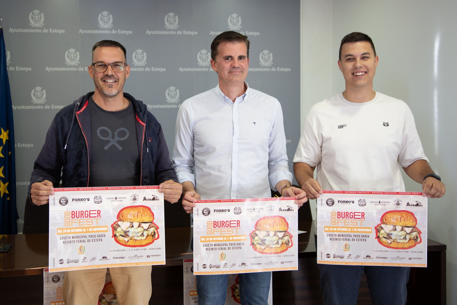 Tres hombres de pie sostienen carteles del Good Burger Fest ante un fondo con el emblema del Ayuntamiento de Estepa.