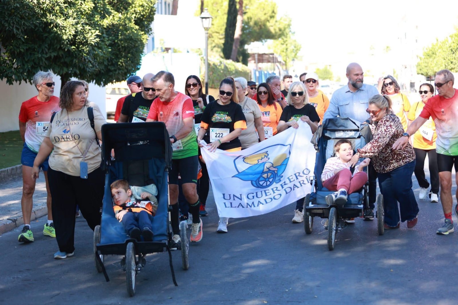 Un grupo de personas con dorsales corre por una calle portando una pancarta y empujando dos carritos con niños.