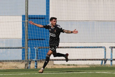 Un futbolista con camiseta negra de lunares blancos corre con los brazos abiertos delante de una portería con red.