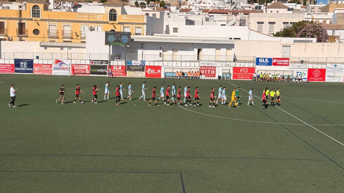 Dos equipos de fútbol con árbitros caminan en fila sobre un campo de césped artificial con vallas publicitarias y edificios blancos al fondo.