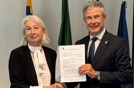 Un hombre y una mujer con traje sostienen un documento escrito junto a tres banderas.