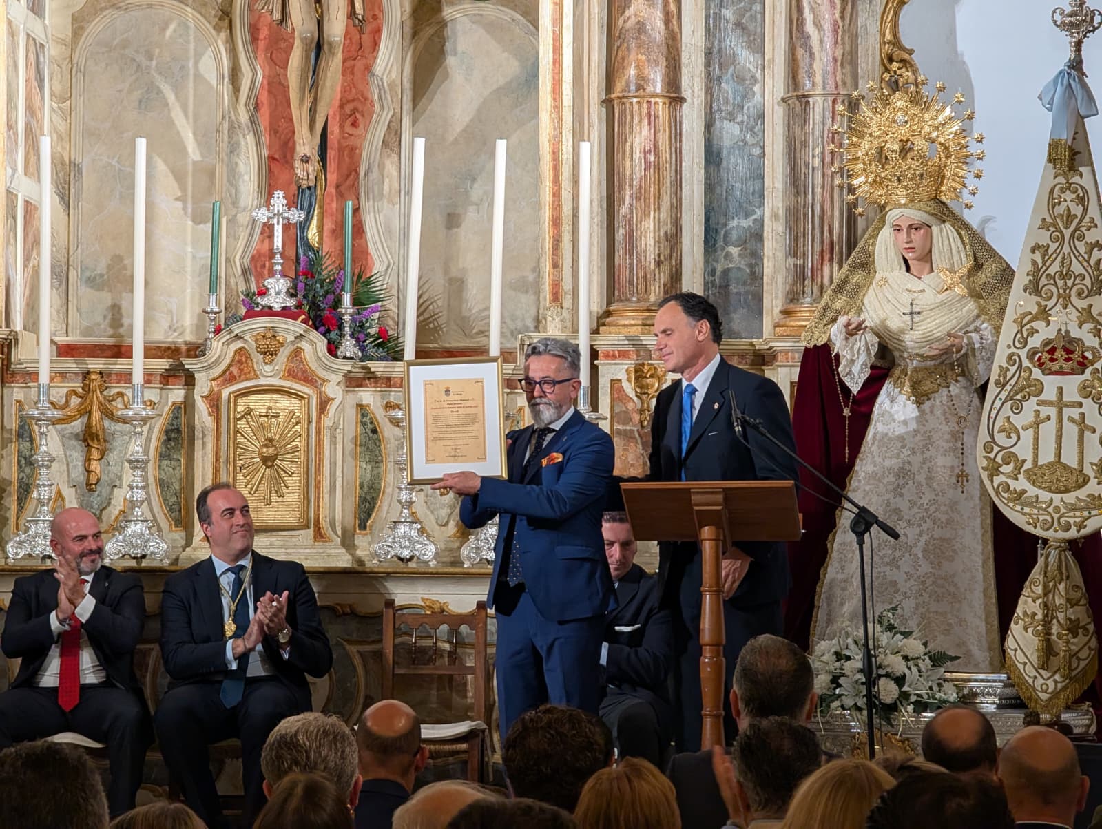 Un hombre con gafas y traje azul sostiene un diploma junto a un atril, ante un altar con una imagen vestida y público sentado.
