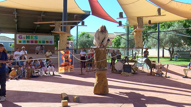 Un artista subido a un tronco de madera actúa ante un público sentado bajo toldos en una plaza.