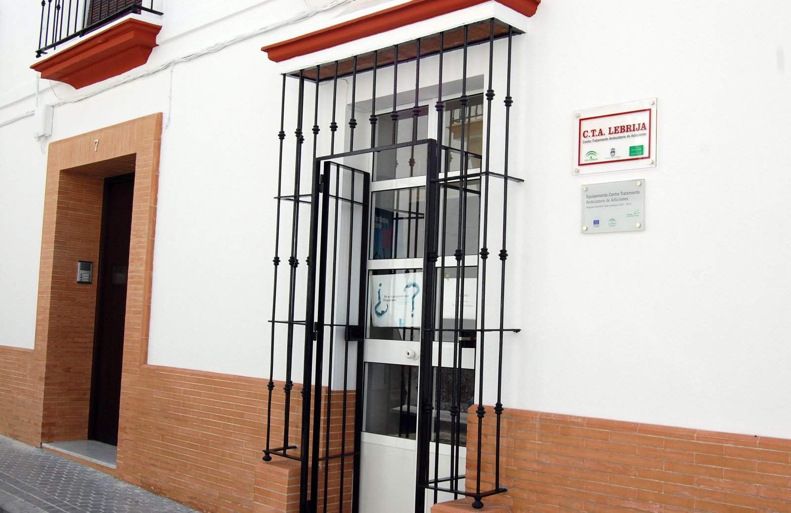 Fachada blanca con zócalo de ladrillo, una puerta con reja negra y dos placas junto a ella.