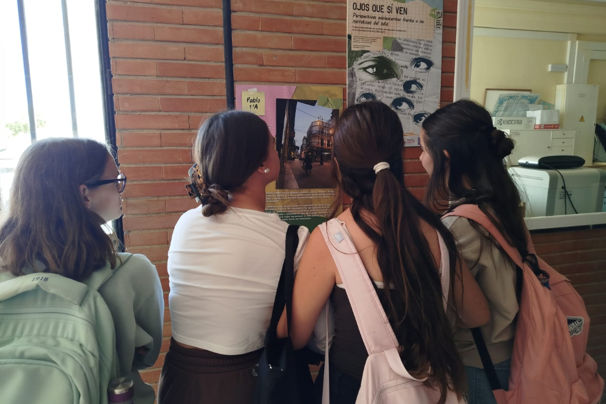 Cuatro jóvenes de espaldas con mochilas miran carteles y un mural con ojos en una pared de ladrillo.