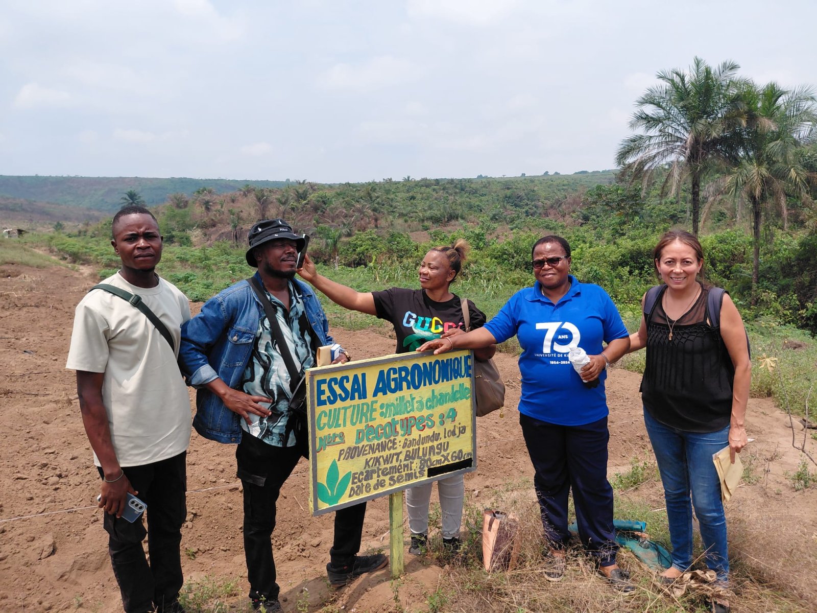 Cinco personas posan junto a un cartel de ensayo agronómico en un campo de tierra con vegetación y palmeras al fondo.