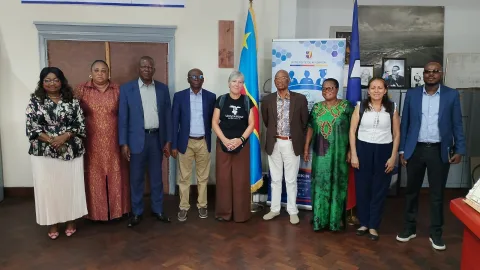 Nueve personas posan de pie en fila en una sala, junto a una bandera azul y amarilla y un cartel con el texto Université de Kinshasa.