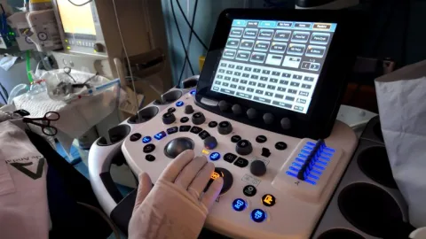 Una mano enguantada maneja un ecógrafo con pantalla táctil de menús y botones, en una sala con equipo médico al fondo.