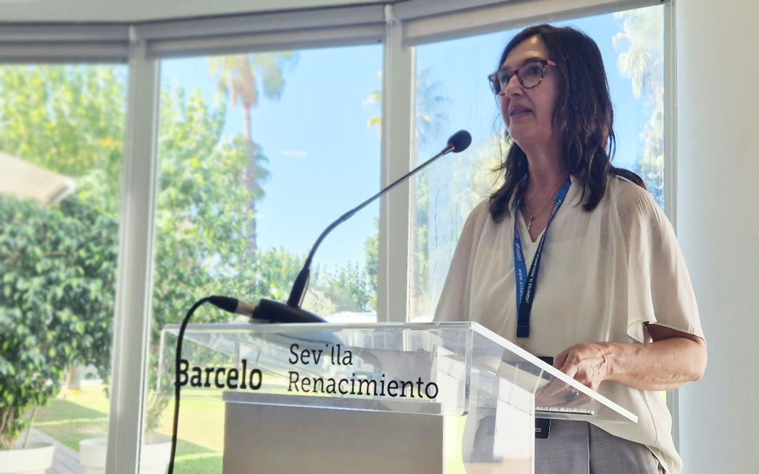Una mujer con gafas y blusa clara habla ante un micrófono tras un atril transparente con el texto Barceló Sevilla Renacimiento, junto a una ventana.