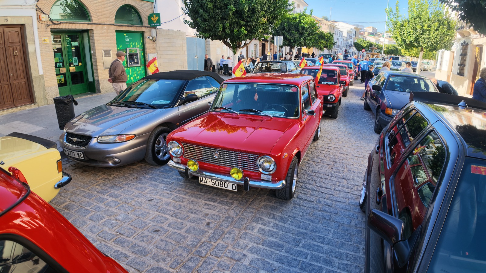 Coches clásicos aparcados en fila en una calle adoquinada, con un turismo rojo en primer plano y personas alrededor.