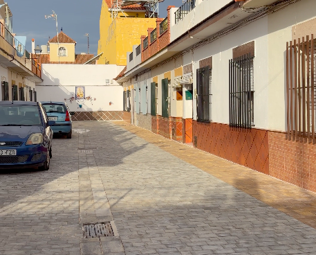 Calle estrecha pavimentada con adoquines, casas blancas con rejas y zócalo de ladrillo, y dos coches aparcados a la izquierda.