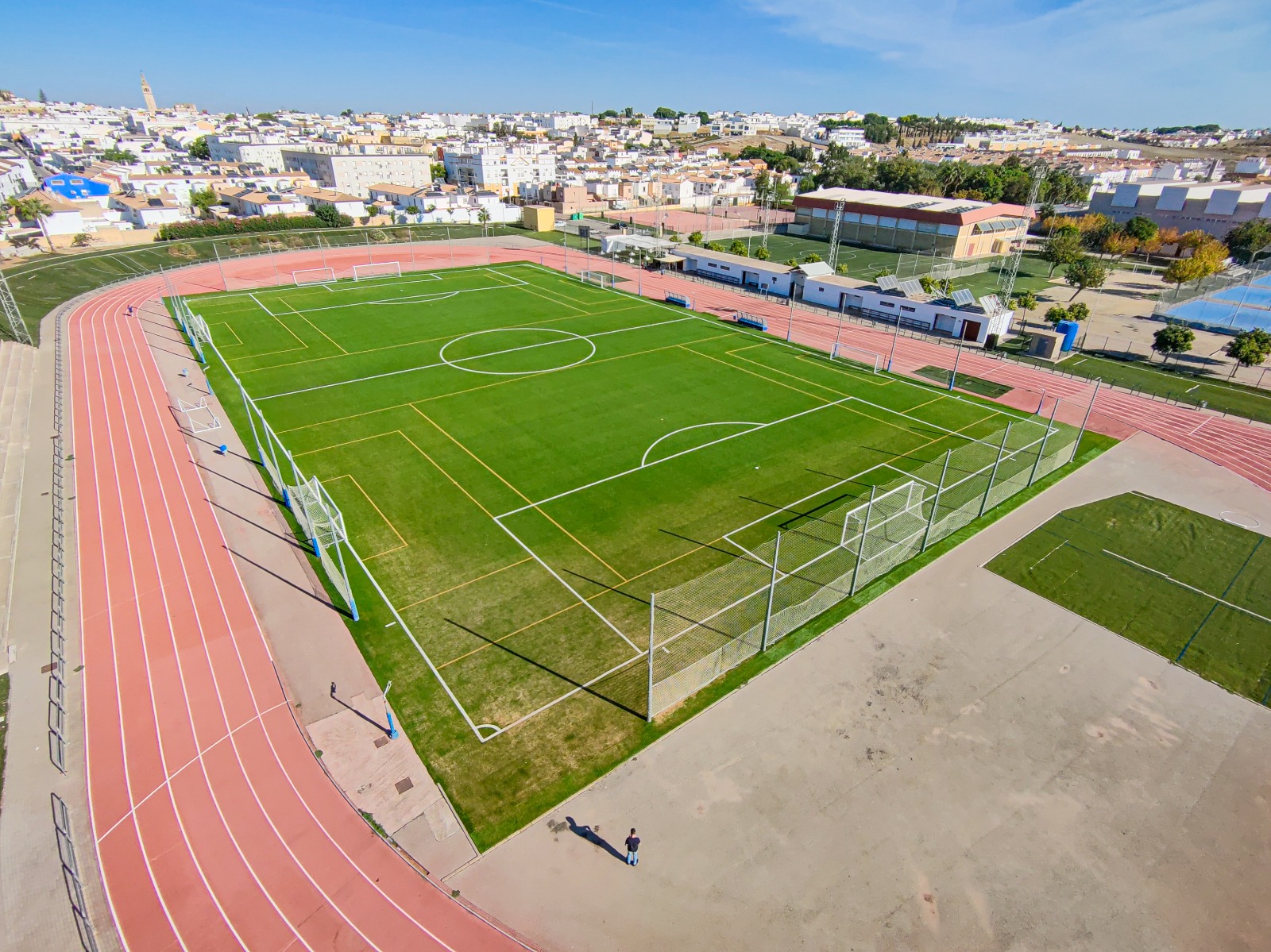 Campo de fútbol de césped verde con pistas de atletismo rojas alrededor y una ciudad blanca al fondo.