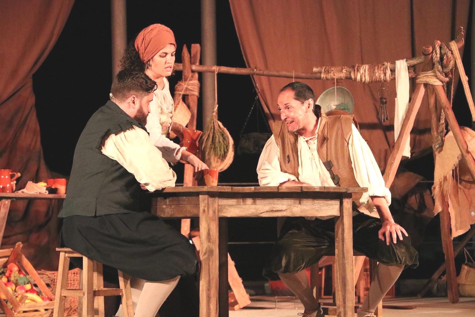 Tres actores con ropa de época conversan alrededor de una mesa de madera en un escenario teatral con decorado rústico.