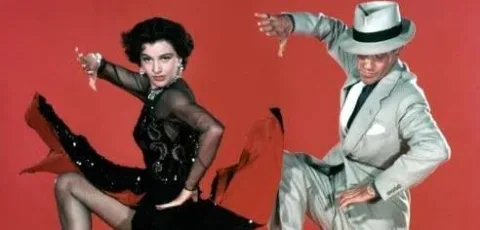 Un hombre con traje gris y sombrero y una mujer con vestido negro de lentejuelas bailan sobre fondo rojo.