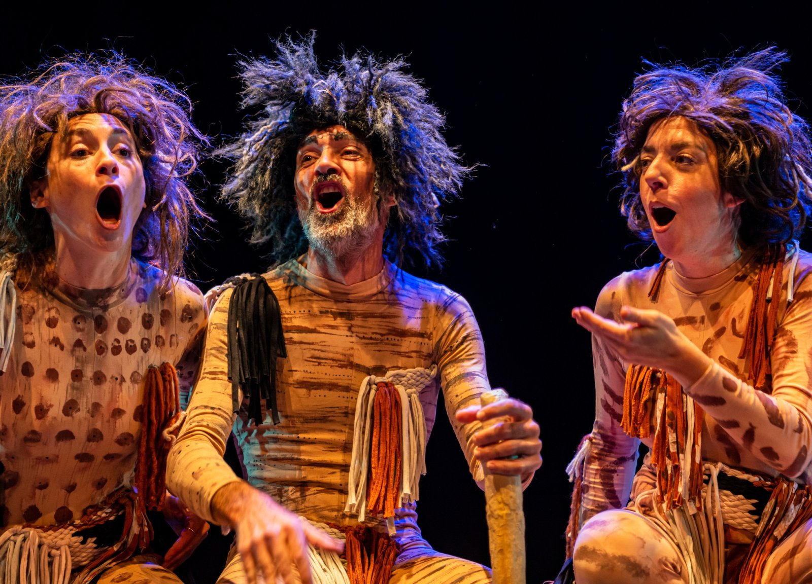 Tres actores caracterizados como personajes prehistóricos, con pelucas desgreñadas y ropas estampadas, cantan con la boca abierta sobre un escenario oscuro.